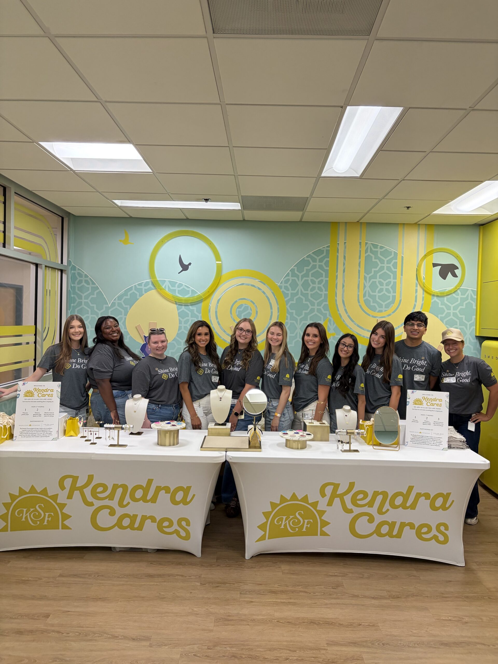 A group of ten volunteers in gray 'Shine Bright, Do Good' t-shirts stand behind two tables set up for a Kendra Cares jewelry-making event. The tables, covered in white spandex covers with yellow 'Kendra Cares' and KCF sunburst logos, display necklaces, bracelets, earrings, and mirrors on gold stands. Signs on the tables explain how the jewelry-making activity works. The group poses in front of a teal and yellow mural featuring birds and geometric circle patterns.